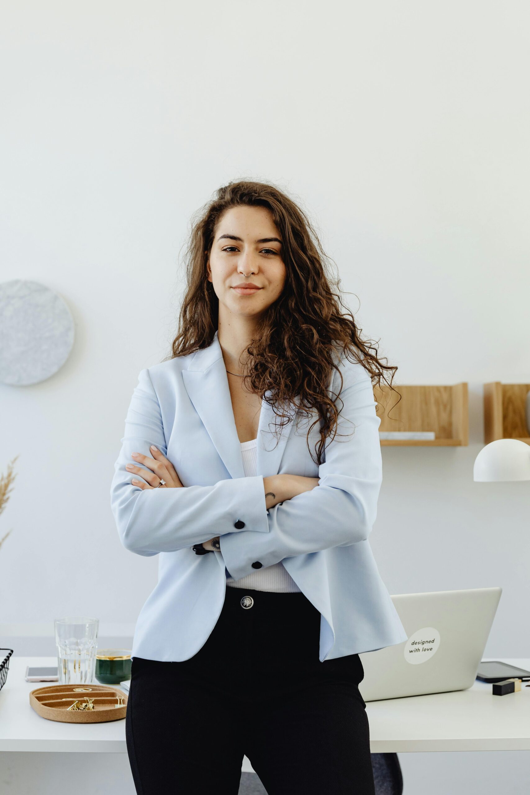 Professional woman in a white blazer standing confidently in an office with arms crossed.